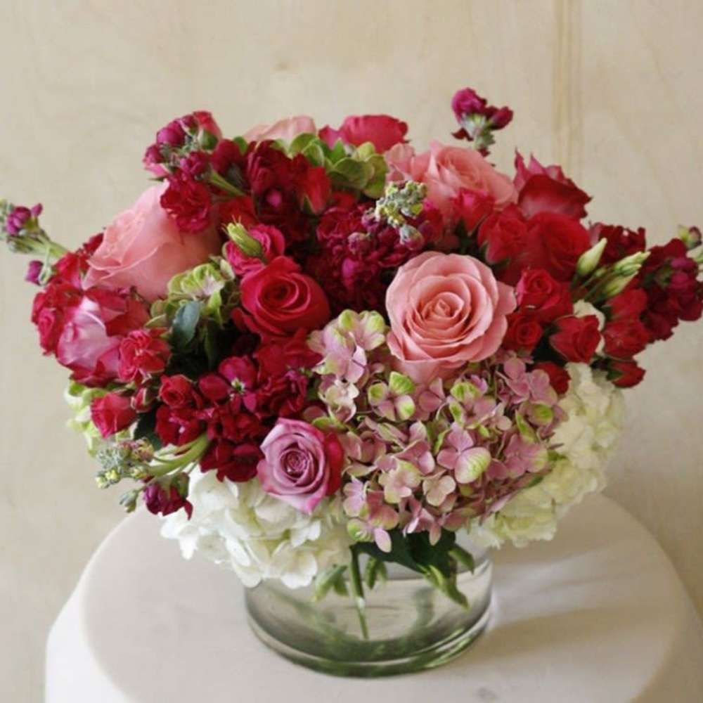 Round arrangement of pink and red roses with hydrangeas and stock in a clear glass cylinder vase