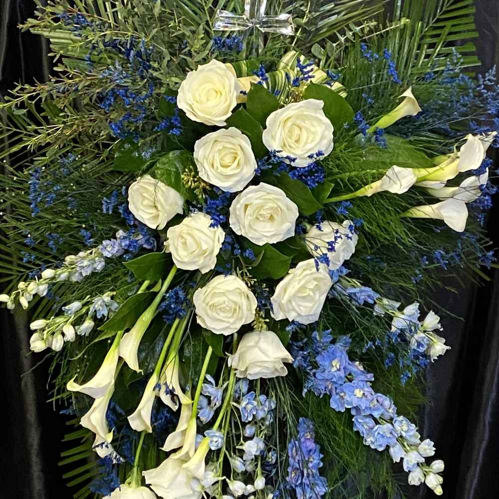 Standing spray of white roses and calla lilies with blue flowers and a glass cross.