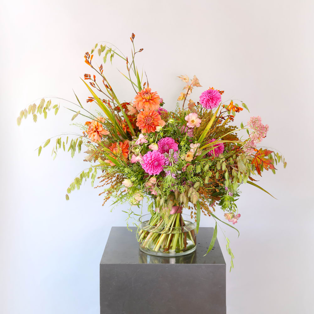 Bouquet of orange and pink dahlias with mixed wildflowers and grasses in a clear glass vase.