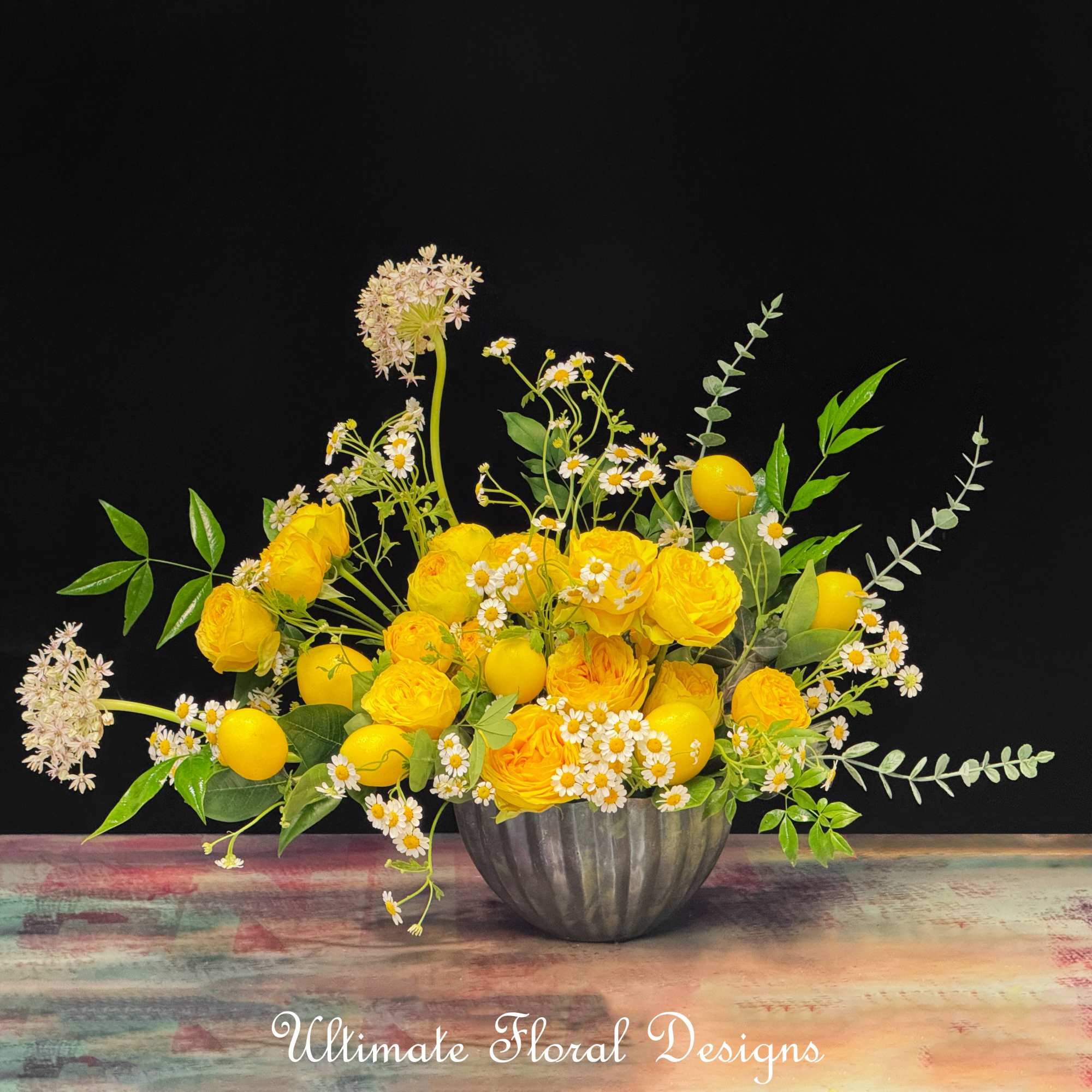 Yellow roses and daisies arranged in a metal bowl with lemon accents