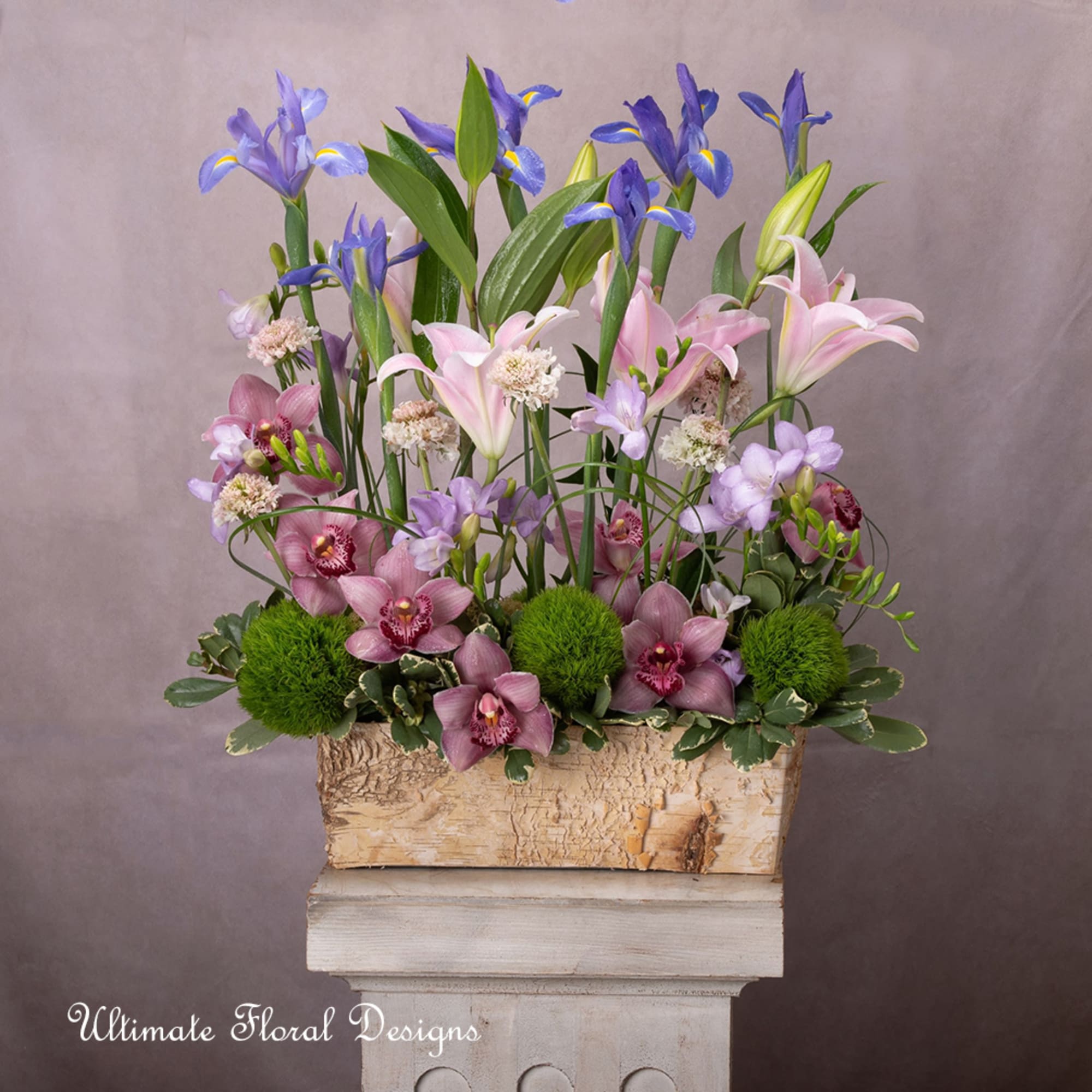 Tall floral arrangement with pink lilies, purple orchids, and green pom-pom blooms in a wooden box
