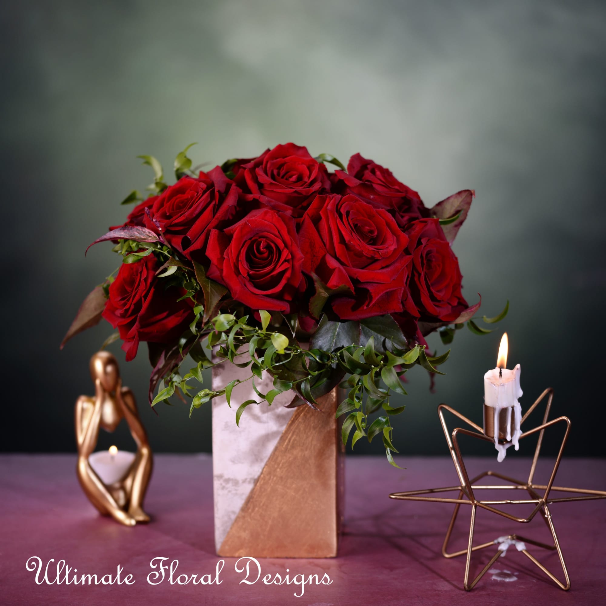 Red roses in a square vase with greenery and candles nearby
