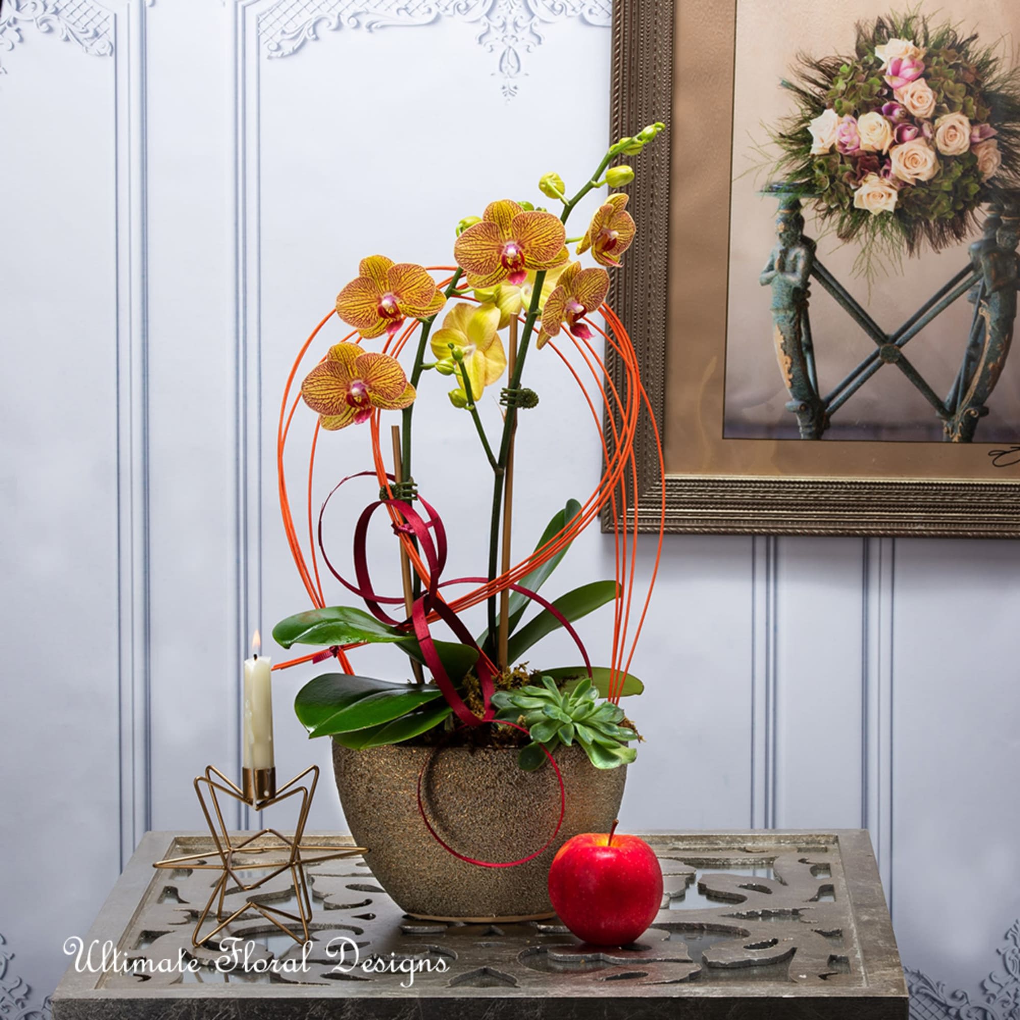 Orchid arrangement in a textured pot with ribbon loops, a candle, and a red apple