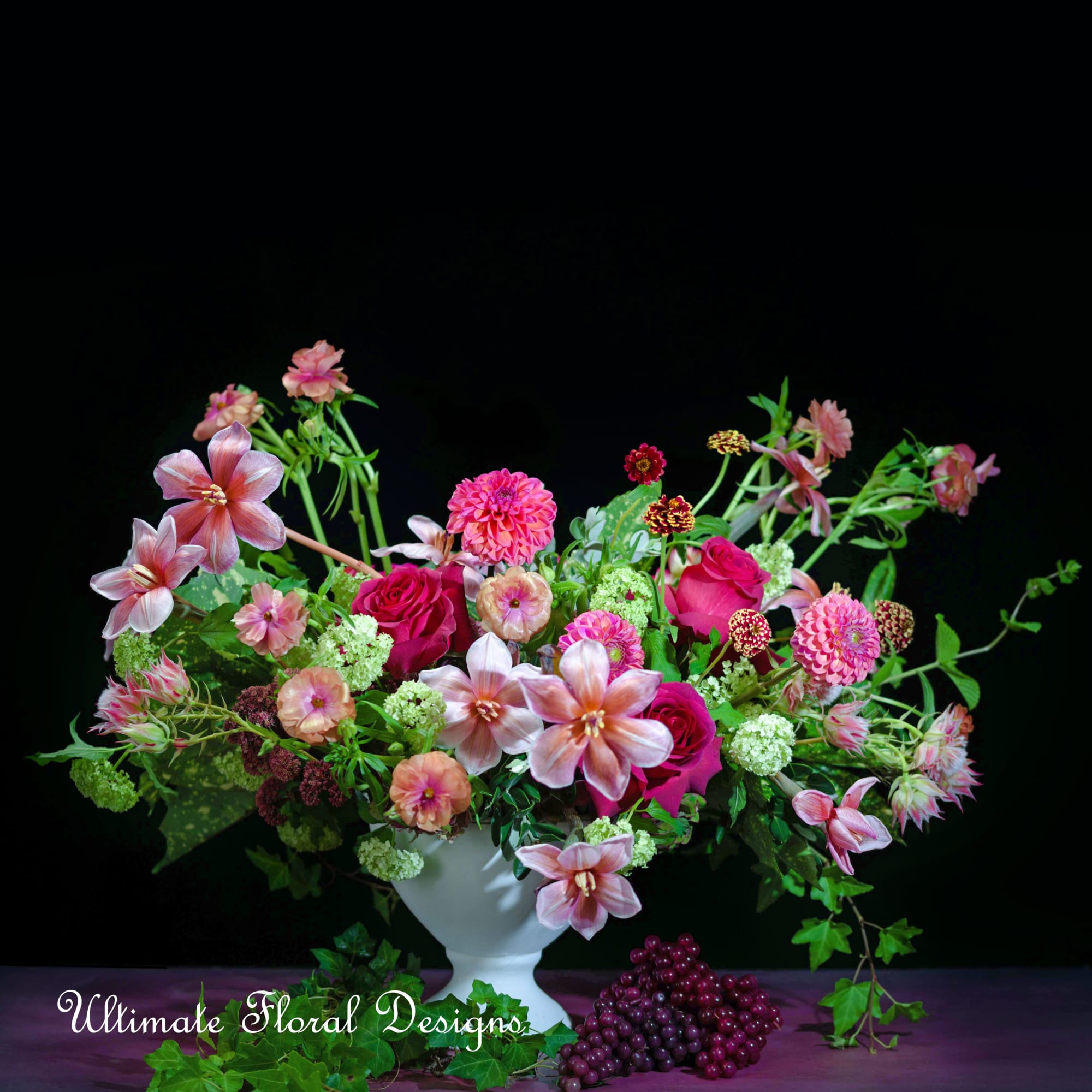 Pink and magenta floral arrangement in a white vase with grapes at the base