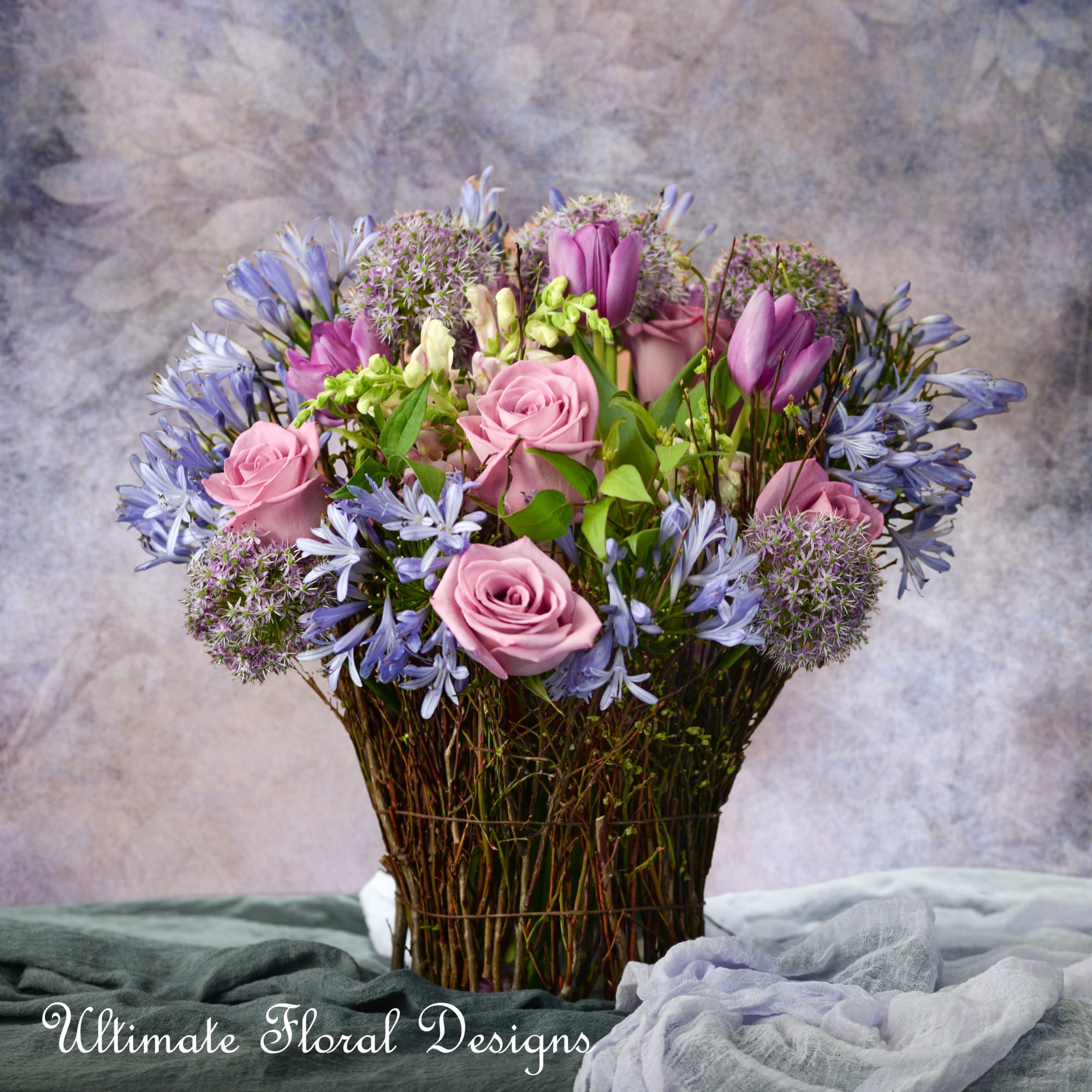 Lavender and pink flower arrangement with roses and tulips in a woven twig basket-style vase.