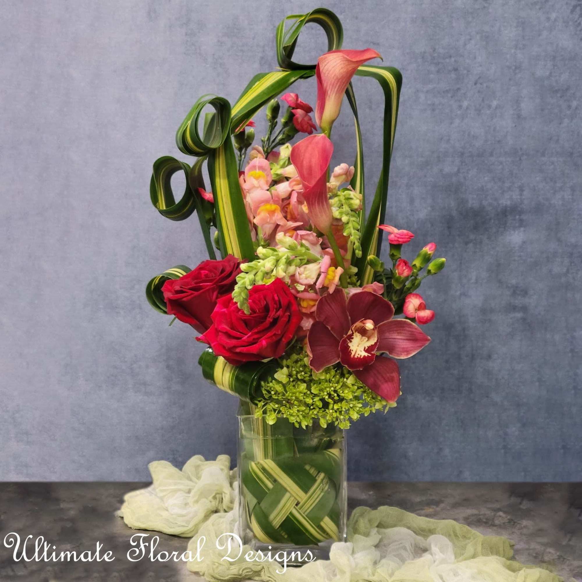 Tall floral arrangement with red roses, pink orchids, and curled green leaves in a glass vase