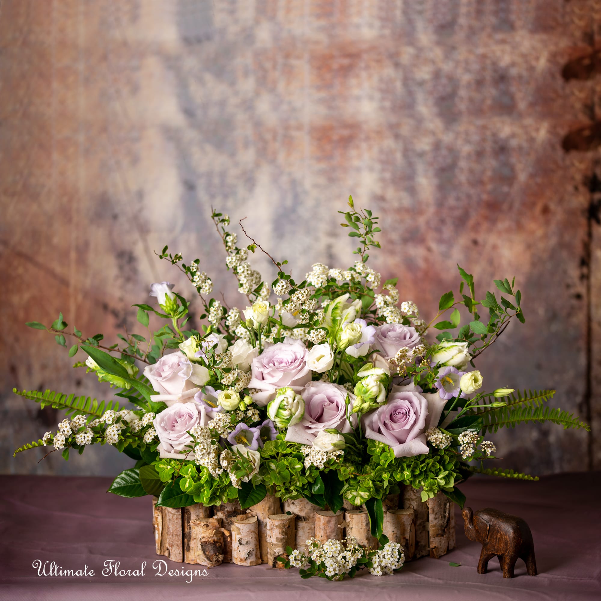 Low arrangement of lavender roses and white blooms in a rustic birch container with a small wooden elephant.