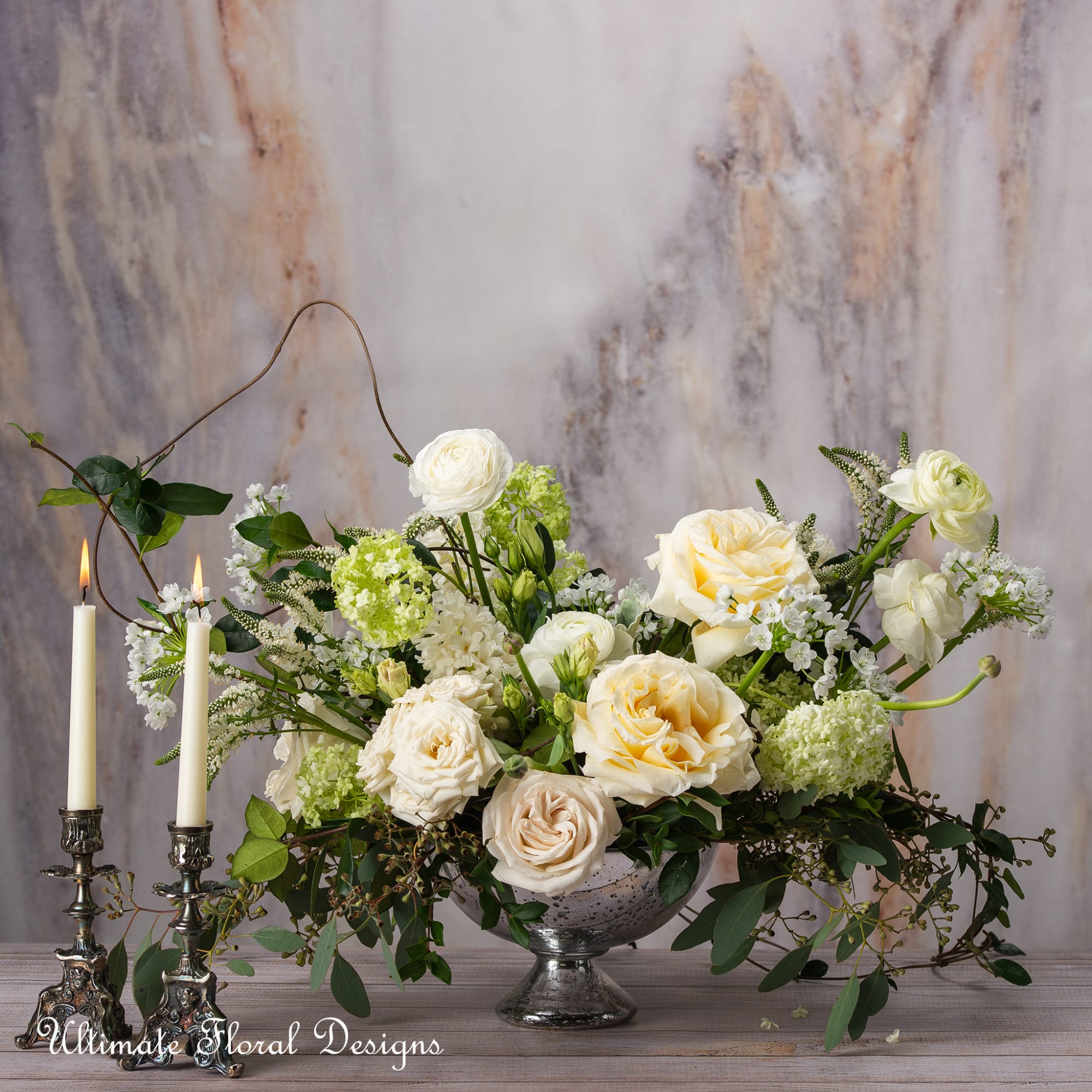 Cream and white floral arrangement in a silver bowl beside two lit candles