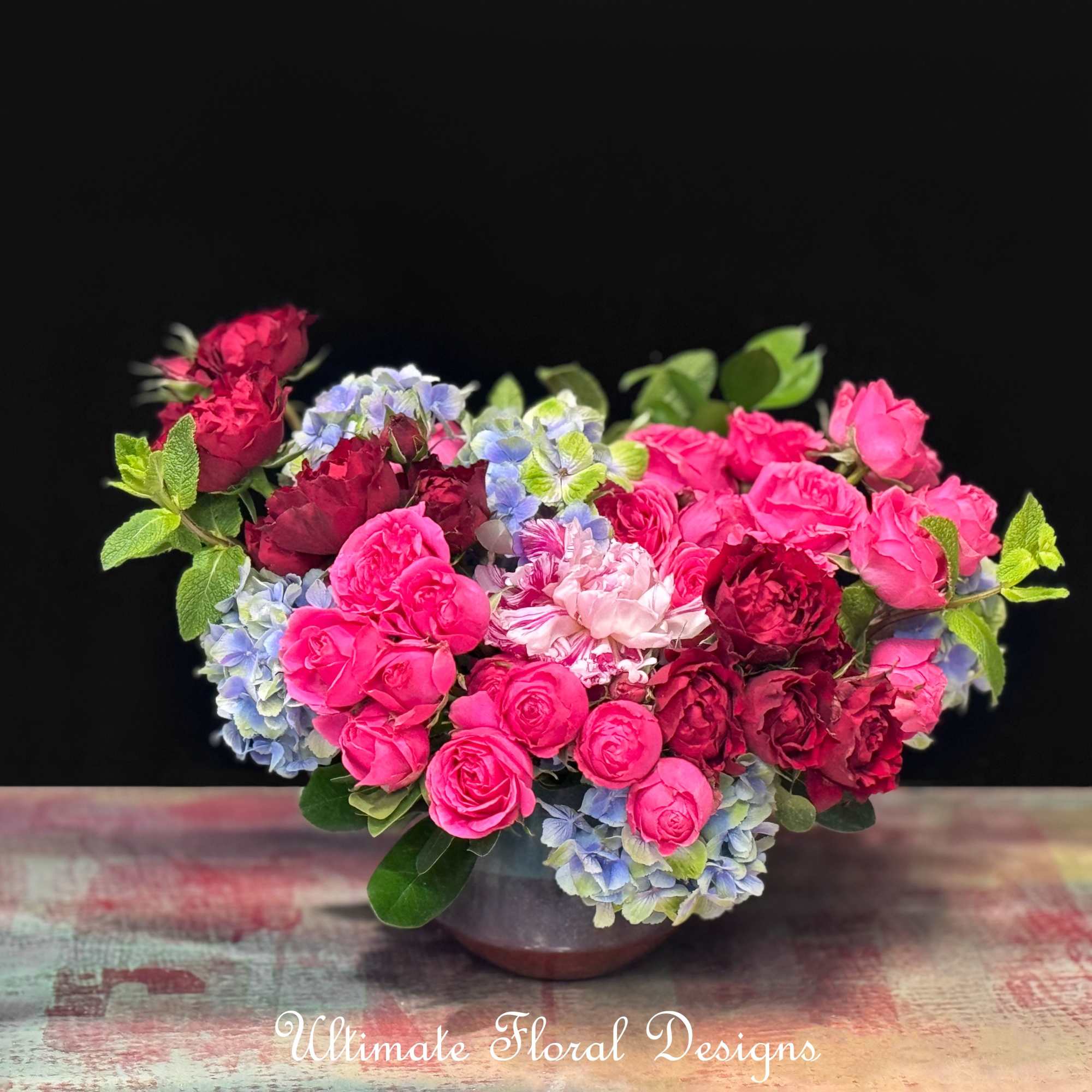 Pink and burgundy roses arranged with blue hydrangeas in a low vase