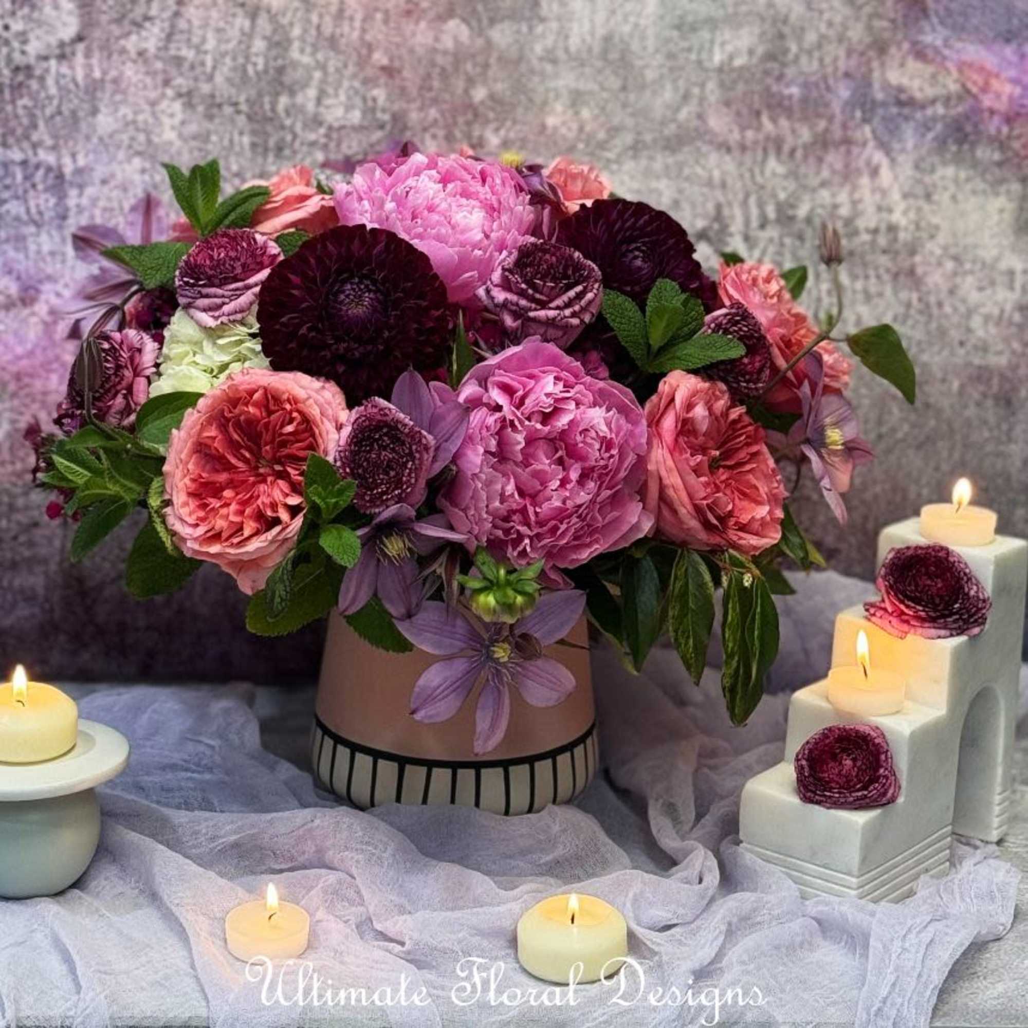 Pink and burgundy floral arrangement in a round box with lit candles nearby.