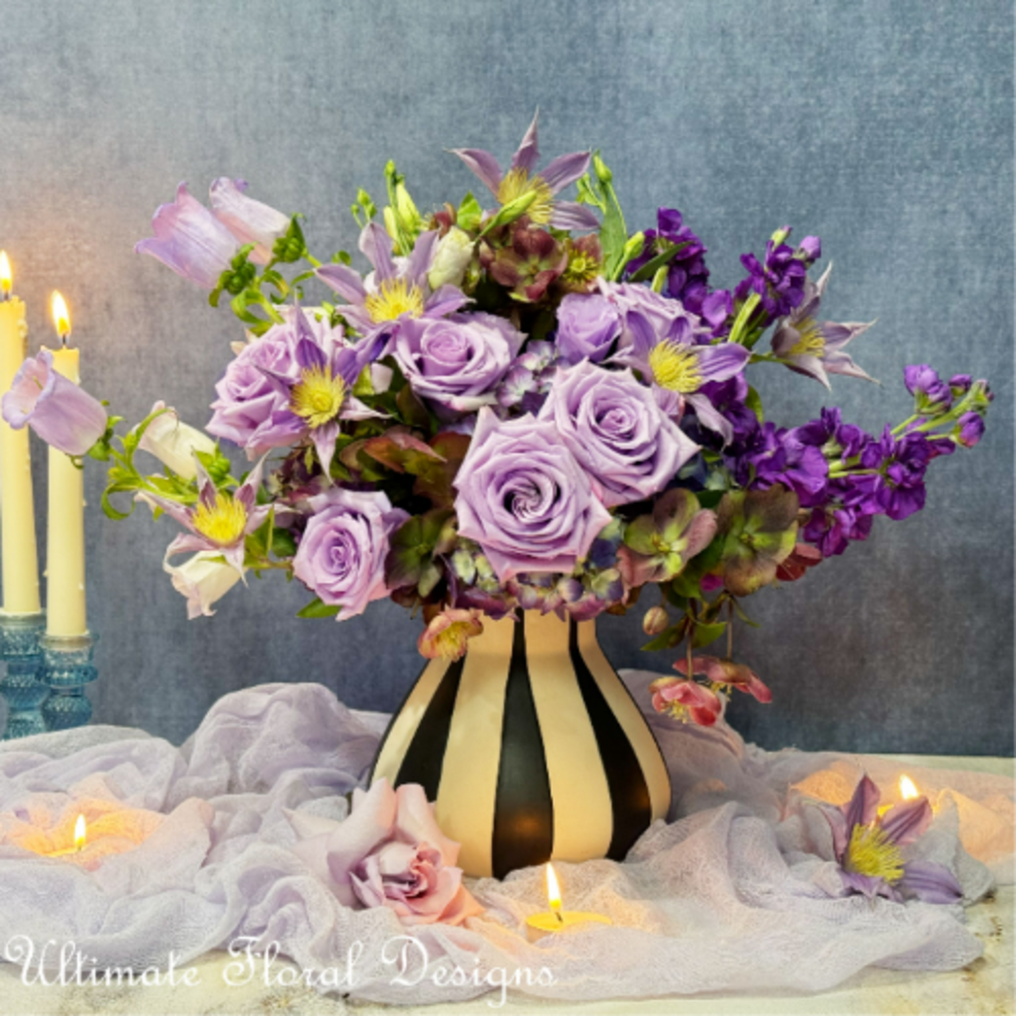 Lavender rose arrangement in a striped vase with purple flowers and candles