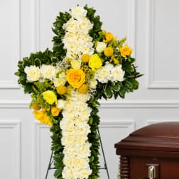 White and yellow floral cross arrangement on a stand beside a casket