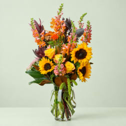 Tall mixed bouquet with sunflowers, yellow roses, and pink snapdragons in a glass vase