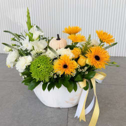 White and yellow floral arrangement in a white bowl with a small pumpkin accent