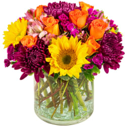 Mixed bouquet of sunflowers, orange roses, and purple mums in a glass vase