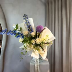 Bouquet of cream roses, blue flowers, and purple accents in white wrap