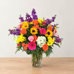 Colorful mixed bouquet in a clear glass vase with sunflowers, roses, and gerbera daisies