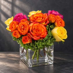 Mixed roses in orange, yellow, and pink arranged in a clear square vase