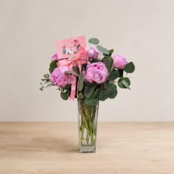 Pink peony bouquet in a clear glass vase with a ribbon and card