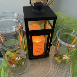 Two glass candle holders flank a black lantern with a memorial message.