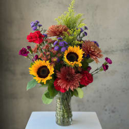 Mixed bouquet with sunflowers, chrysanthemums, and purple accent flowers in a glass vase.