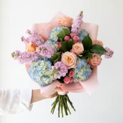 Handheld bouquet of pink roses, blue hydrangeas, and lavender blooms wrapped in pink paper