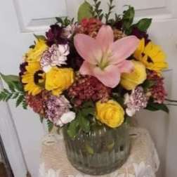 Mixed bouquet with a pink lily, yellow roses, and purple flowers in a glass vase