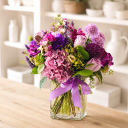 Mixed pink and purple bouquet in a clear square vase with a lavender ribbon