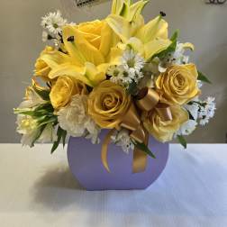 Yellow roses and lilies arranged in a lavender box with a gold ribbon