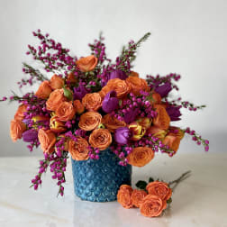 Orange and purple bouquet in a blue textured vase with a few roses beside it