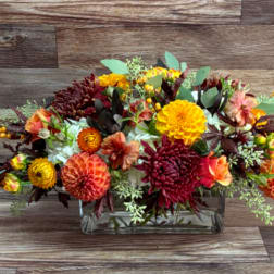Low floral arrangement in a clear rectangular vase with orange, yellow, burgundy, and white blooms