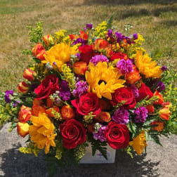 Bright mixed bouquet of red roses, yellow blooms, and purple flowers in a white container