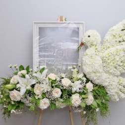 White floral funeral tribute with roses, lilies, and a dove-shaped spray on an easel