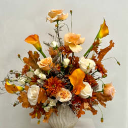 Autumn floral arrangement with peach roses, white calla lilies, and orange mums in a white vase.