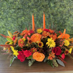 Low floral centerpiece with orange, red, and yellow blooms and two tall orange candles