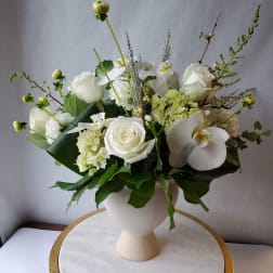 White arrangement of roses, hydrangea, and orchids in a white pedestal vase on a marble stand