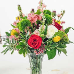 Mixed bouquet of red roses, yellow tulips, white blooms, and pink flowers in a clear glass vase