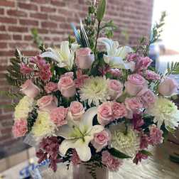 Arrangement of pink roses, carnations, and white lilies in a textured vase on a wooden table.