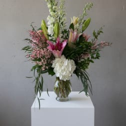 Tall arrangement of pink lilies, white hydrangea, and white stock in a clear glass vase