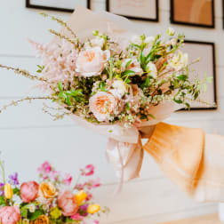 Hand-tied bouquet of peach and cream flowers wrapped in blush paper with a ribbon bow.