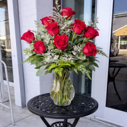 Arrangement of long-stem red roses with white filler flowers in a clear glass vase on a black metal table.