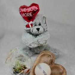 Plush teddy bear holding a red heart balloon beside a small plant globe and soap gift