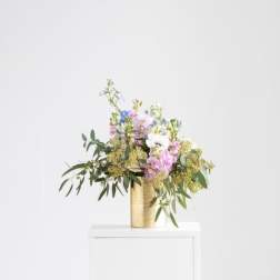 Pastel mixed flower arrangement in a gold cylinder vase on a white pedestal