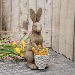 Rabbit figurine holding a basket of carrots beside flowers