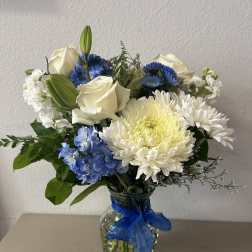 White roses and blue flowers arranged in a glass vase with a blue ribbon.