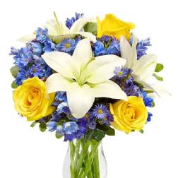 Clear vase of white lilies, yellow roses, and blue flowers in a rounded mixed bouquet