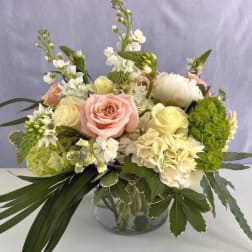 Low arrangement of pale pink roses, white blooms, and lush greenery in a clear glass bowl vase
