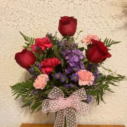 Bouquet of red roses, purple flowers, and pink carnations in a glass vase with a ribbon