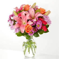 Bouquet of pink lilies, orange roses, and purple daisies in a glass vase
