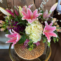 Arrangement of pink lilies, white hydrangea, and purple flowers in a vase with woven cover on a glass table.
